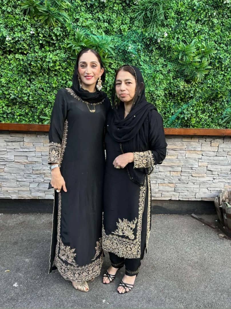 Two women in elegant black qameez with intricate designs, standing against a greenery backdrop, showcasing modest fashion.