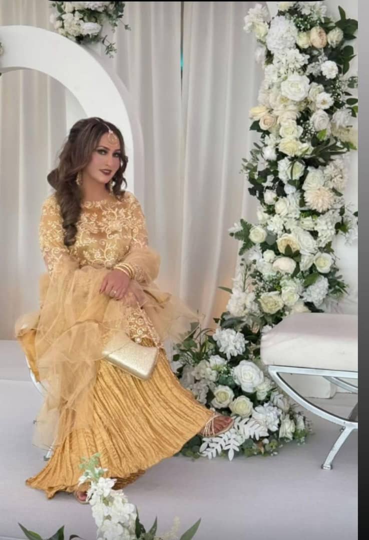 Elegant woman in a golden beige net qameez surrounded by floral decor, showcasing sophistication and style.