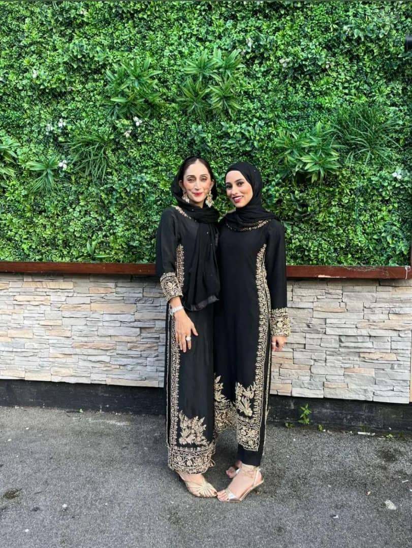 Two women in elegant black qameez with gold embroidery posed against a green wall, showcasing a blend of tradition and style.
