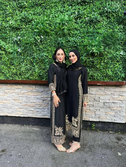 Two women in elegant black qameez with gold embroidery posed against a green wall, showcasing a blend of tradition and style.