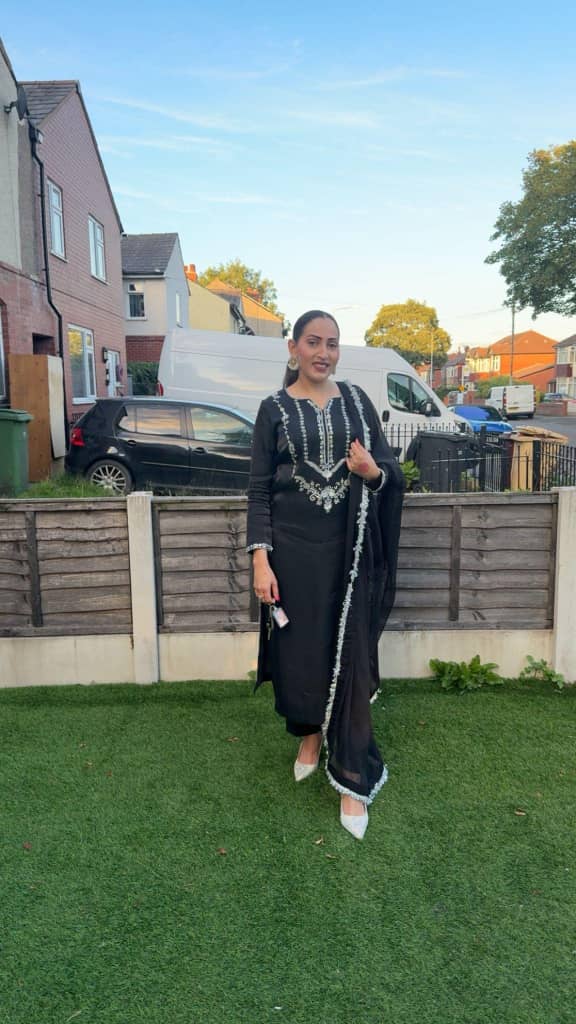 Woman in a black embroidered outfit standing in a garden, showcasing traditional fashion against a suburban background.
