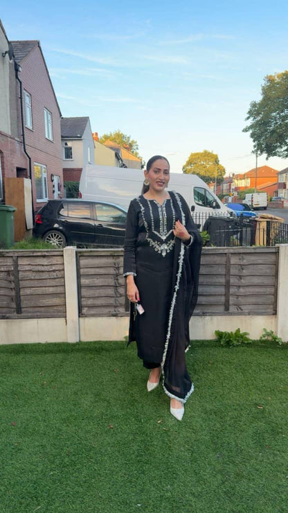 Woman in a black embroidered outfit standing in a garden, showcasing traditional fashion against a suburban background.